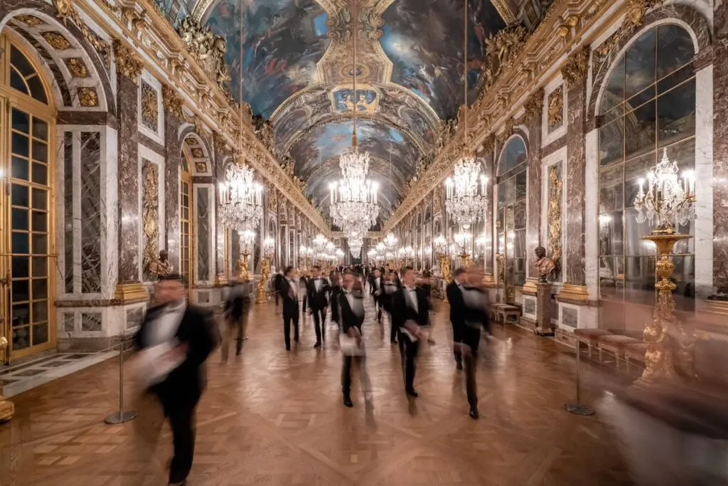 Hommes en smoking traversant la Galerie des Glaces du Château de Versailles lors d'une soirée privée, photographiés en pose lente.