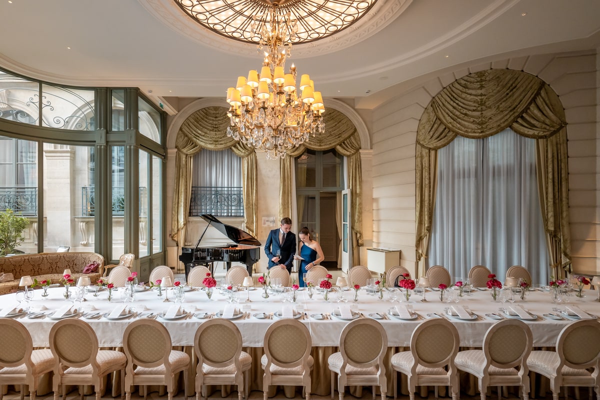 Grande table de banquet dressée dans un salon luxueux du Ritz Paris avec lustre monumental, piano à queue et hauts plafonds.