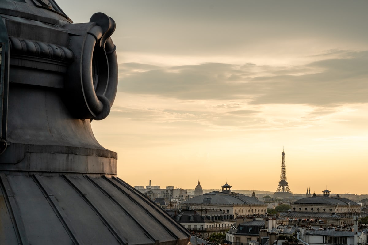 Fenêtre en œil-de-bœuf au premier plan et tour Eiffel au crépuscule vue depuis le rooftop Terraza Mikuna à Paris.